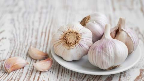 4 garlic bulbs sitting in a white dish and 3 garlic cloves, both sitting on a white timber table